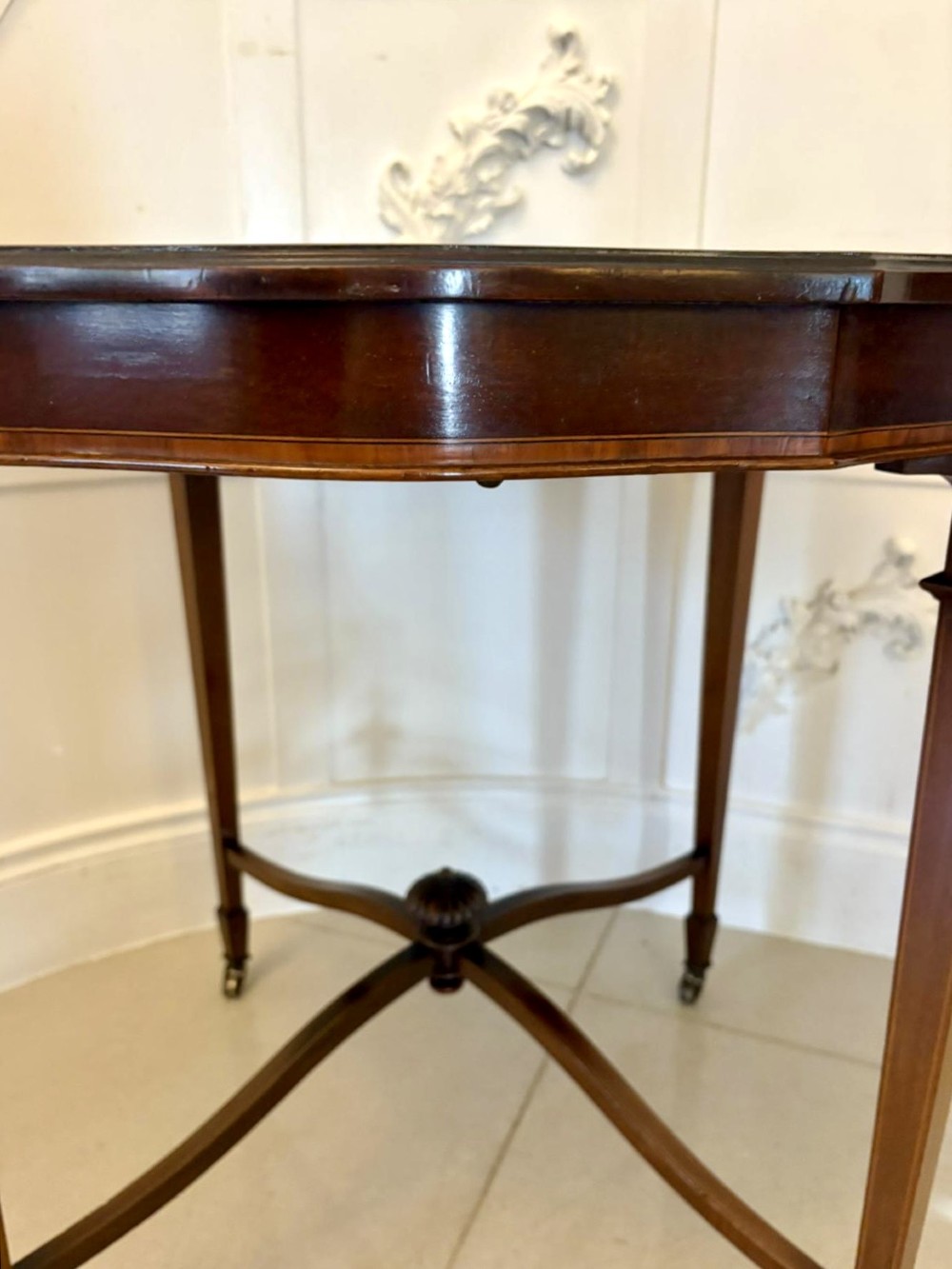 An antique Edwardian polished wood centre table featuring a shaped scalloped top, satinwood inlaid banding, tapered legs with brass castors, and an X-frame stretcher with a central finial. View V