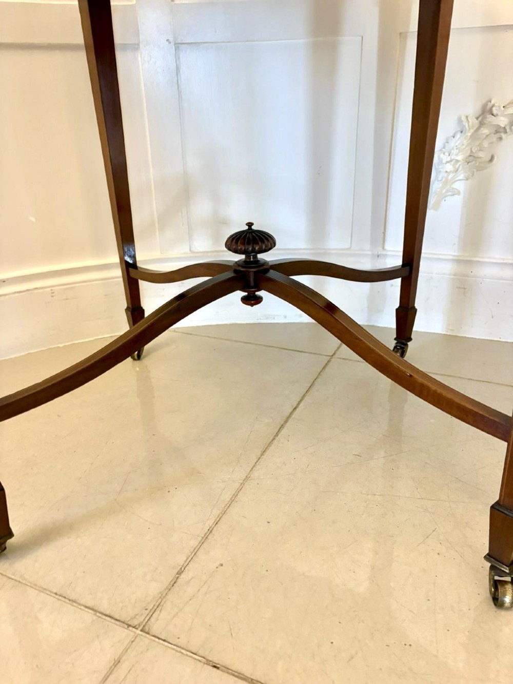 An antique Edwardian polished wood centre table featuring a shaped scalloped top, satinwood inlaid banding, tapered legs with brass castors, and an X-frame stretcher with a central finial. View VI
