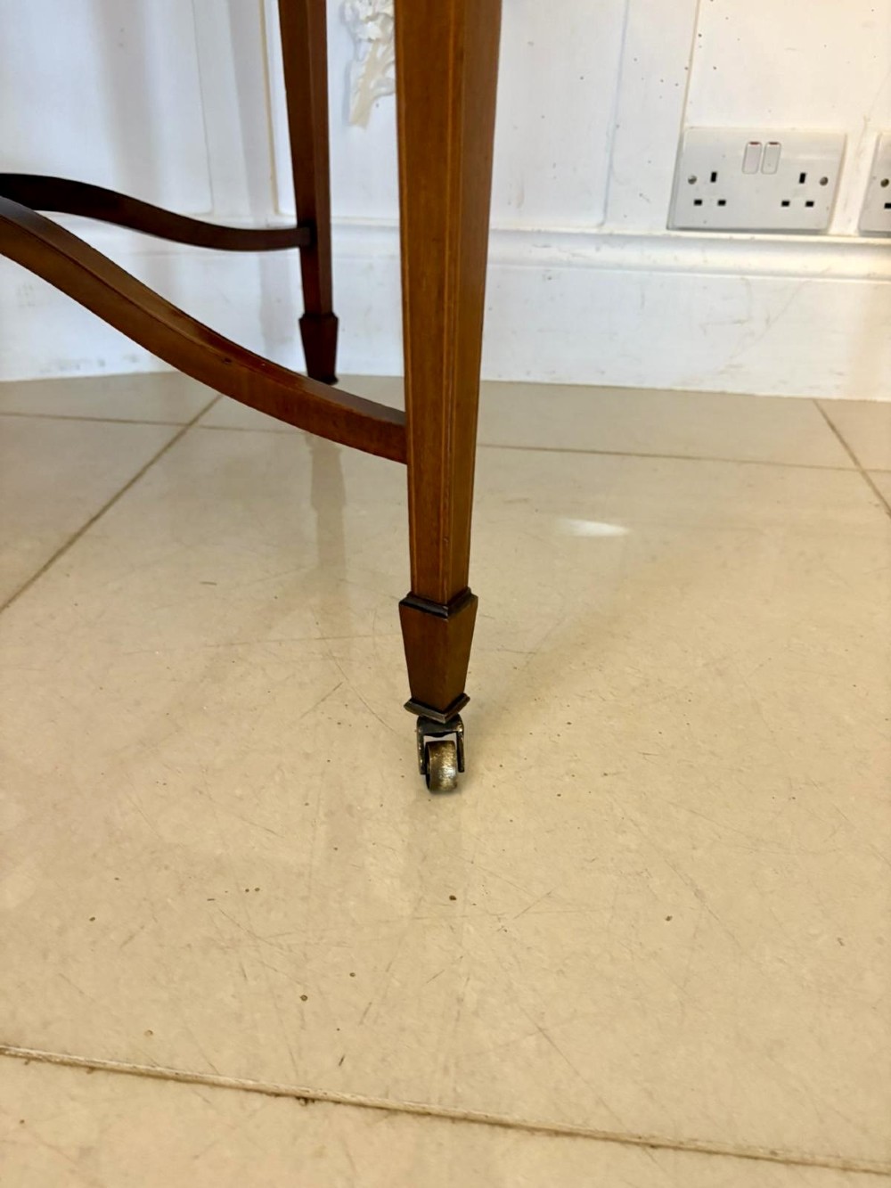 An antique Edwardian polished wood centre table featuring a shaped scalloped top, satinwood inlaid banding, tapered legs with brass castors, and an X-frame stretcher with a central finial. View VII