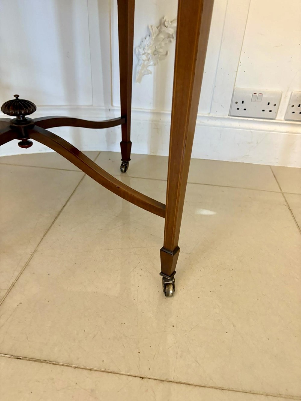 An antique Edwardian polished wood centre table featuring a shaped scalloped top, satinwood inlaid banding, tapered legs with brass castors, and an X-frame stretcher with a central finial. View VIII