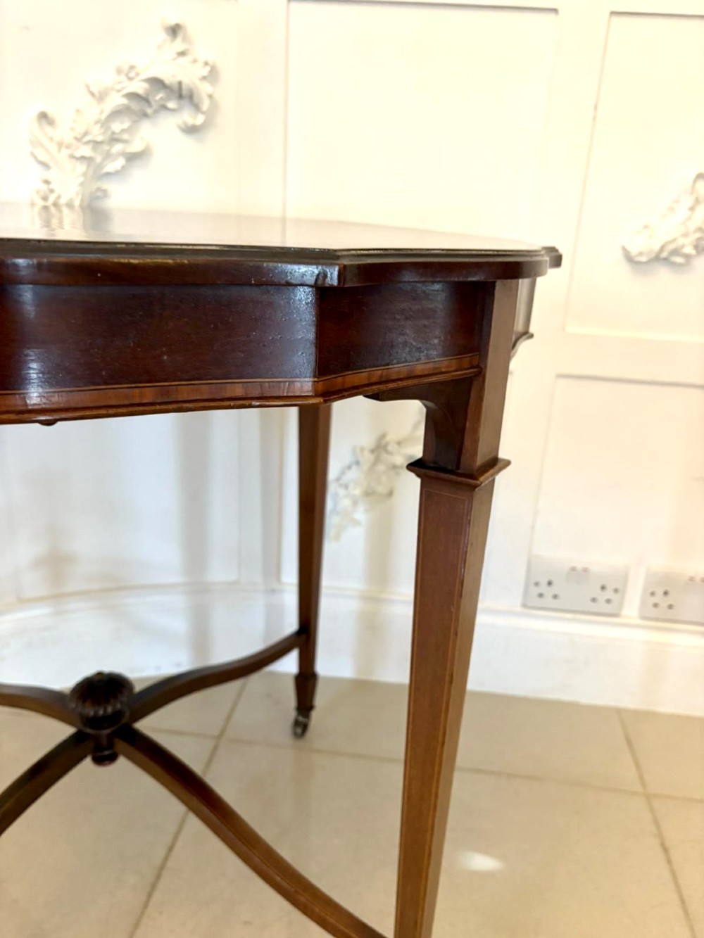 An antique Edwardian polished wood centre table featuring a shaped scalloped top, satinwood inlaid banding, tapered legs with brass castors, and an X-frame stretcher with a central finial. View IX