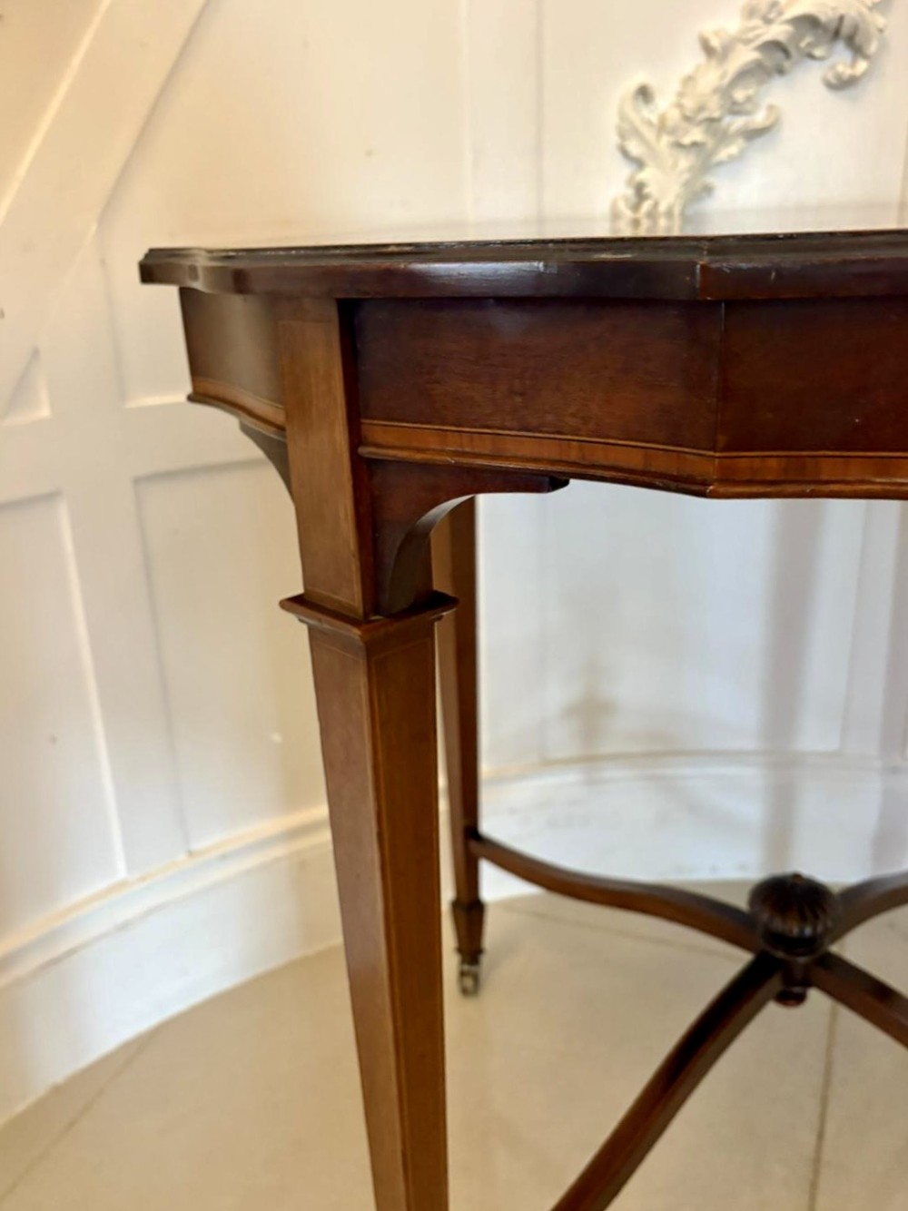 An antique Edwardian polished wood centre table featuring a shaped scalloped top, satinwood inlaid banding, tapered legs with brass castors, and an X-frame stretcher with a central finial. View X