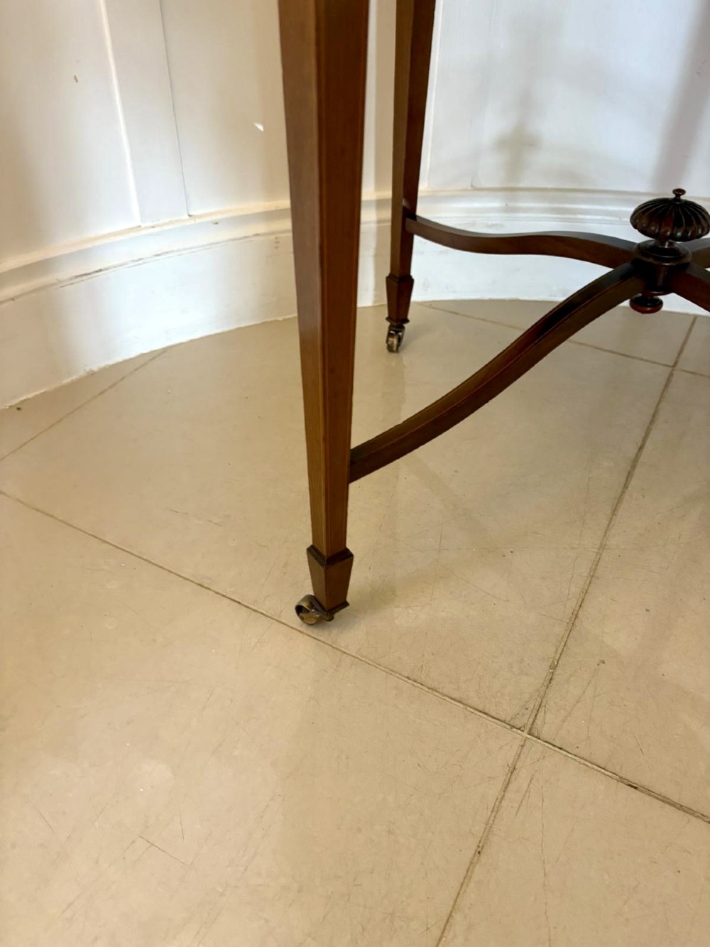 An antique Edwardian polished wood centre table featuring a shaped scalloped top, satinwood inlaid banding, tapered legs with brass castors, and an X-frame stretcher with a central finial. View XI
