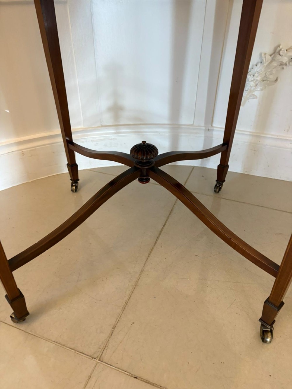 An antique Edwardian polished wood centre table featuring a shaped scalloped top, satinwood inlaid banding, tapered legs with brass castors, and an X-frame stretcher with a central finial. View XII