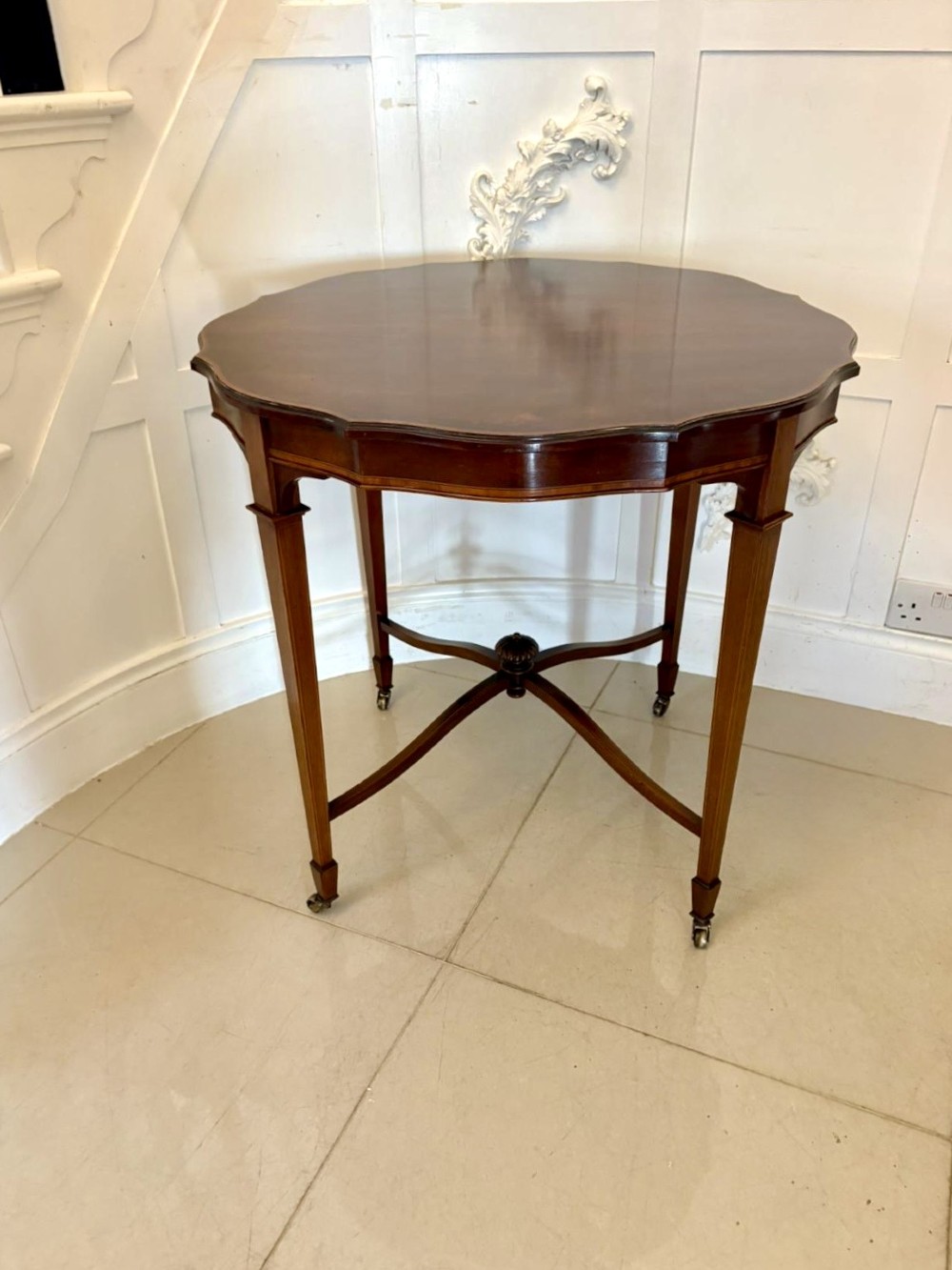 An antique Edwardian polished wood centre table featuring a shaped scalloped top, satinwood inlaid banding, tapered legs with brass castors, and an X-frame stretcher with a central finial.
