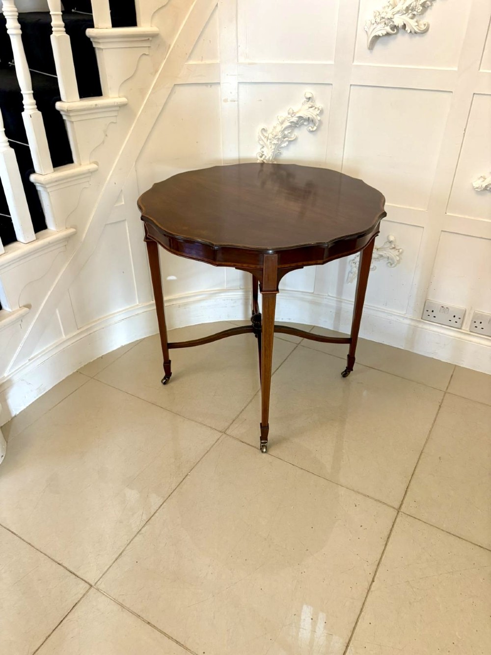 An antique Edwardian polished wood centre table featuring a shaped scalloped top, satinwood inlaid banding, tapered legs with brass castors, and an X-frame stretcher with a central finial. View XIII