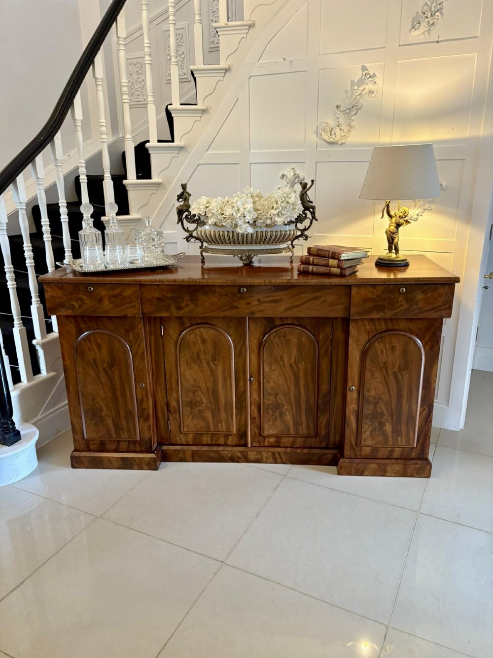 A Victorian figured mahogany sideboard with four arched cupboard doors, three shallow frieze drawers, and a plinth base. View V