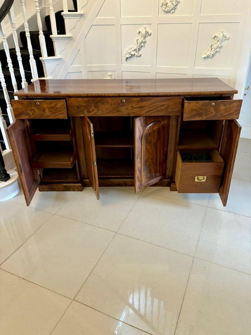 A Victorian figured mahogany sideboard with four arched cupboard doors, three shallow frieze drawers, and a plinth base. View VI