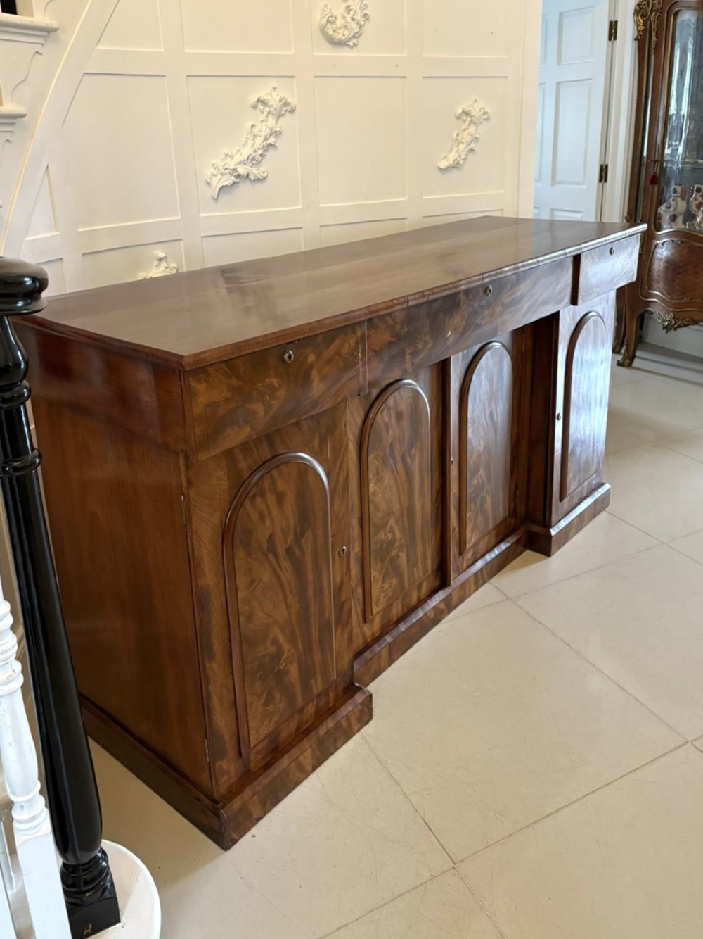 A Victorian figured mahogany sideboard with four arched cupboard doors, three shallow frieze drawers, and a plinth base. View X