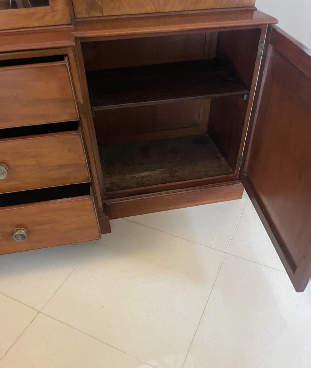 A large George III mahogany breakfront bookcase featuring glazed upper doors topped with a broken swan-neck pediment, set over a base section with drawers and side cupboards. View VI