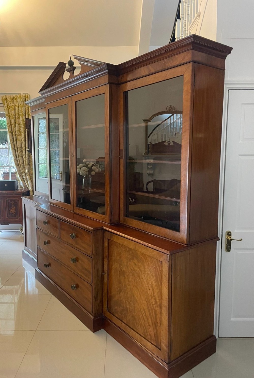 A large George III mahogany breakfront bookcase featuring glazed upper doors topped with a broken swan-neck pediment, set over a base section with drawers and side cupboards. View XI