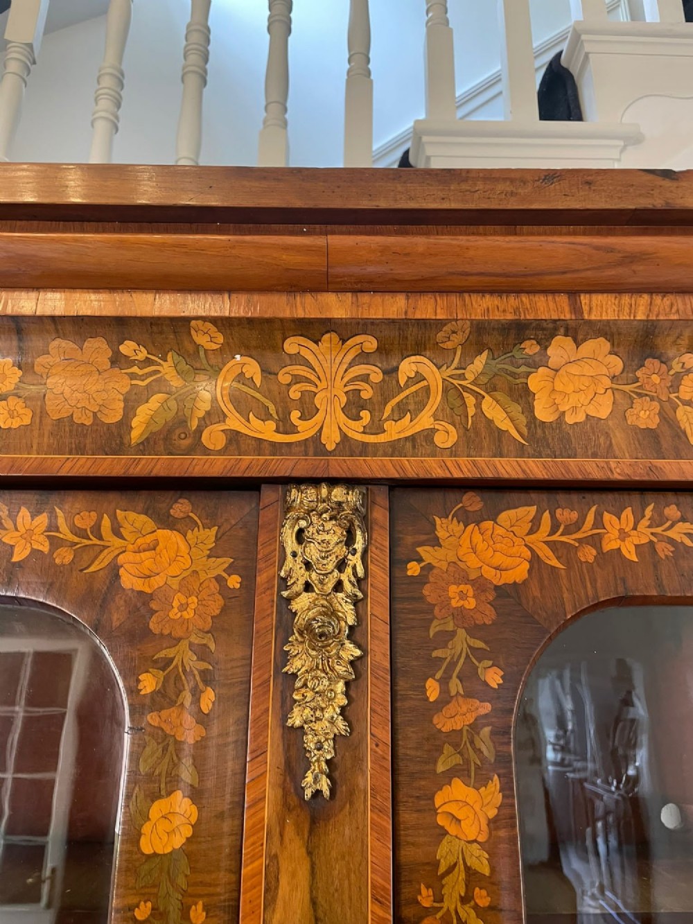 A Victorian two-part burr walnut bookcase cabinet featuring glazed upper doors with floral marquetry inlays and gilt ormolu mounts. View VI
