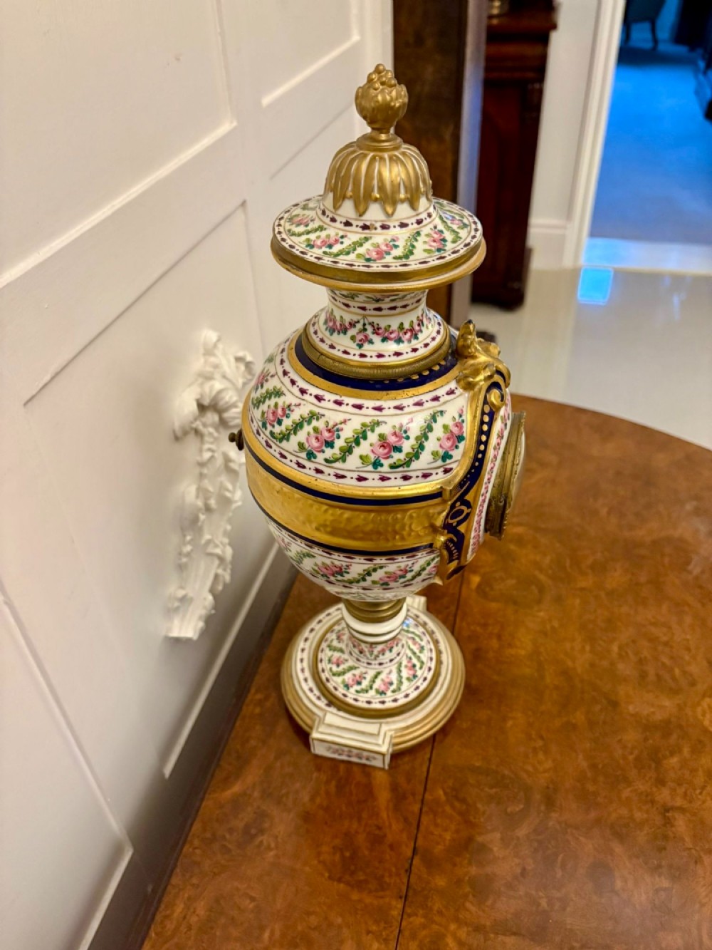 A French hand-painted porcelain urn-shaped mantel clock featuring gold leaf accents, cobalt blue borders, and pink floral garlands on a gilded pedestal base. View II