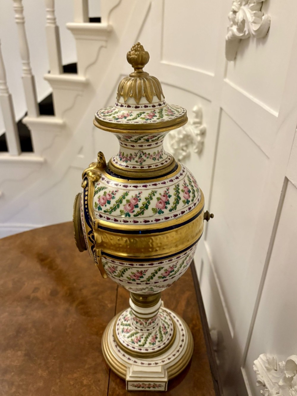 A French hand-painted porcelain urn-shaped mantel clock featuring gold leaf accents, cobalt blue borders, and pink floral garlands on a gilded pedestal base. View III
