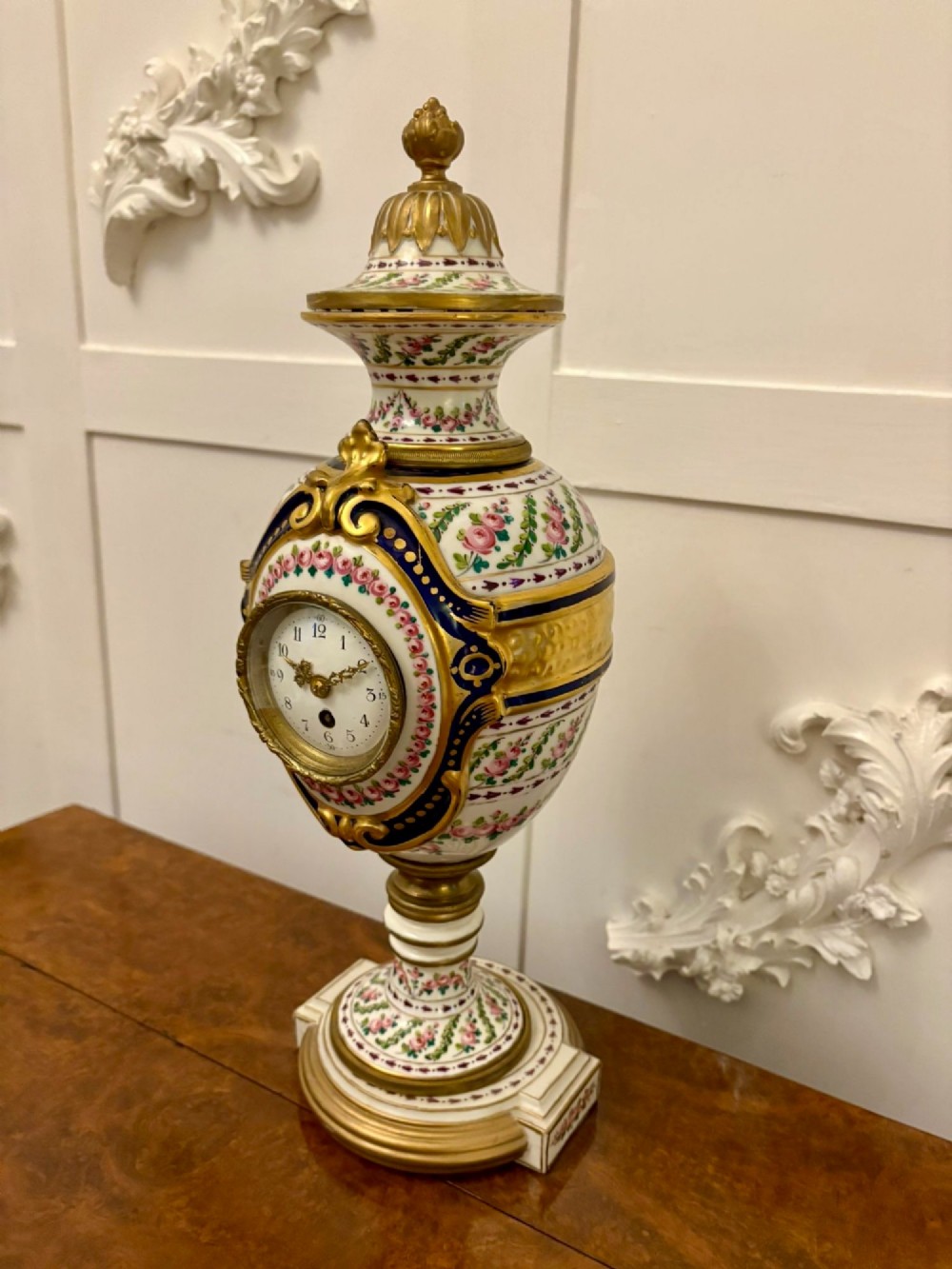 A French hand-painted porcelain urn-shaped mantel clock featuring gold leaf accents, cobalt blue borders, and pink floral garlands on a gilded pedestal base. View IV
