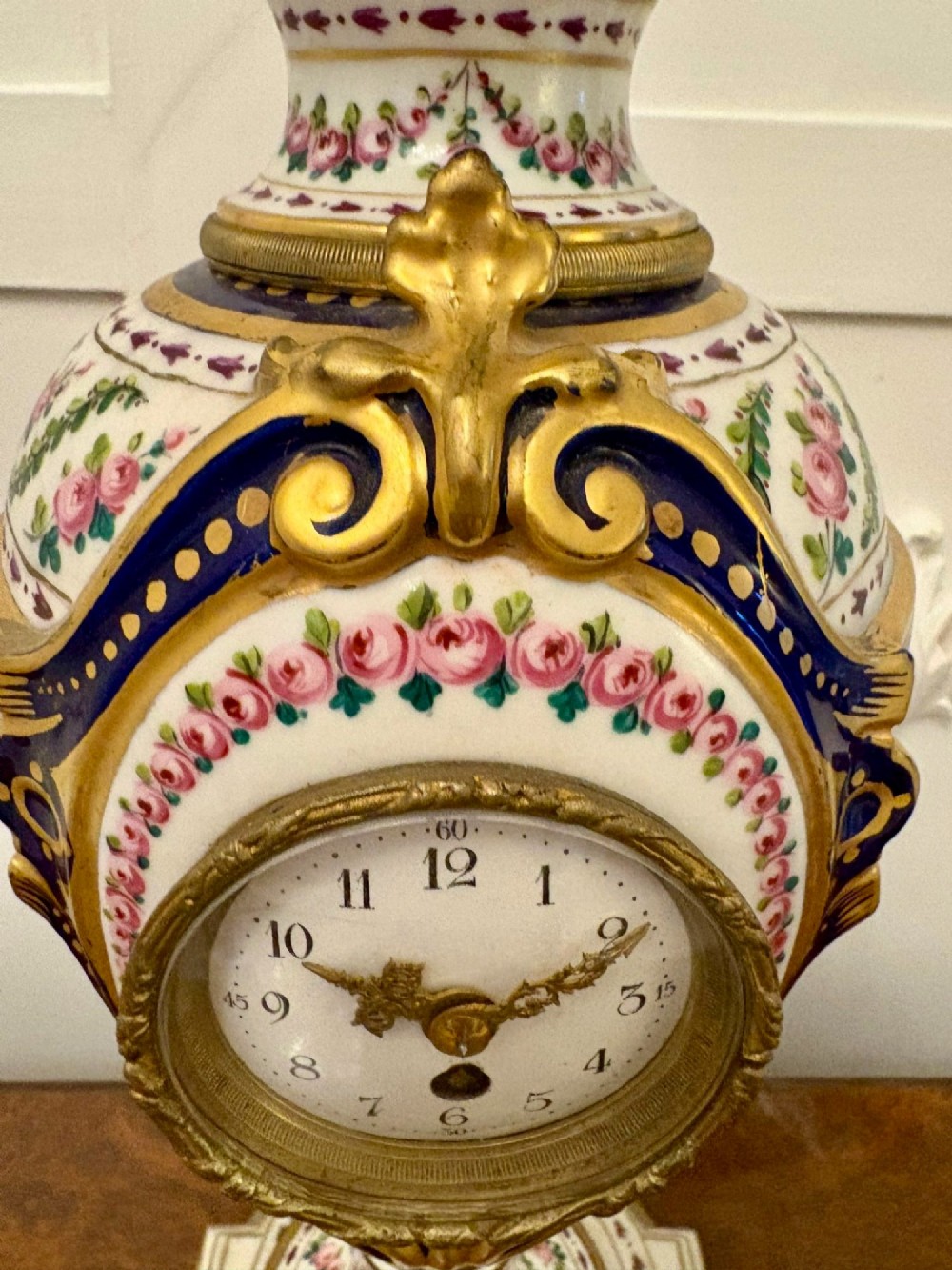 A French hand-painted porcelain urn-shaped mantel clock featuring gold leaf accents, cobalt blue borders, and pink floral garlands on a gilded pedestal base. View V