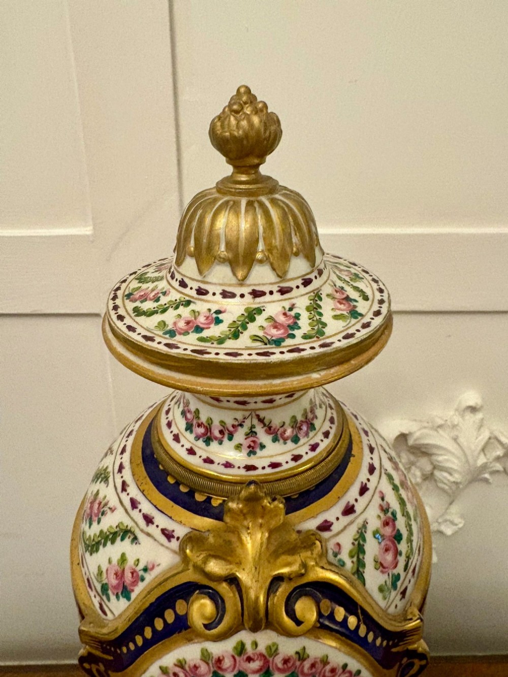 A French hand-painted porcelain urn-shaped mantel clock featuring gold leaf accents, cobalt blue borders, and pink floral garlands on a gilded pedestal base. View VII