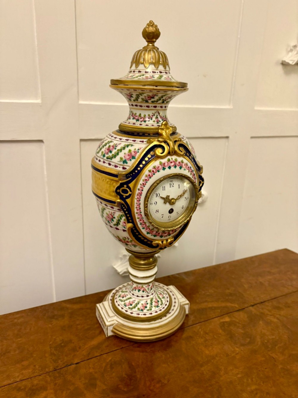 A French hand-painted porcelain urn-shaped mantel clock featuring gold leaf accents, cobalt blue borders, and pink floral garlands on a gilded pedestal base. View VIII