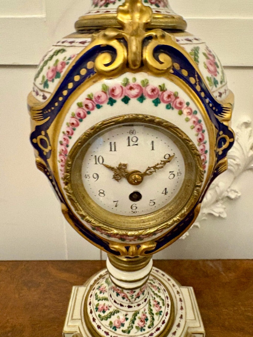 A French hand-painted porcelain urn-shaped mantel clock featuring gold leaf accents, cobalt blue borders, and pink floral garlands on a gilded pedestal base. View IX