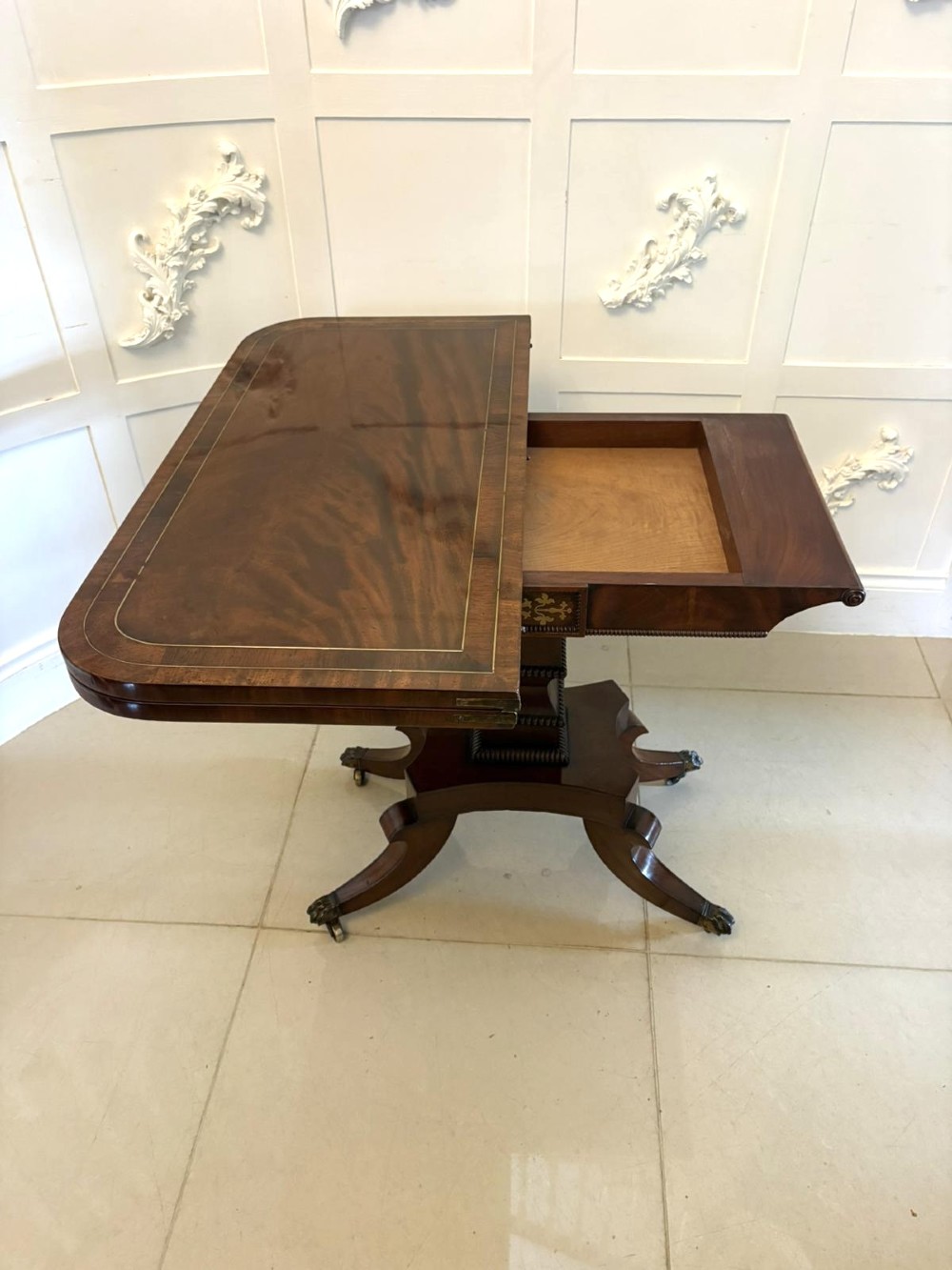 An antique Regency mahogany fold-over card table featuring brass marquetry inlay, a central shaped pedestal, and a splayed base with brass castors. View VII