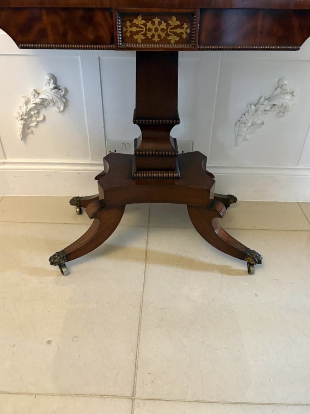 An antique Regency mahogany fold-over card table featuring brass marquetry inlay, a central shaped pedestal, and a splayed base with brass castors. View XII
