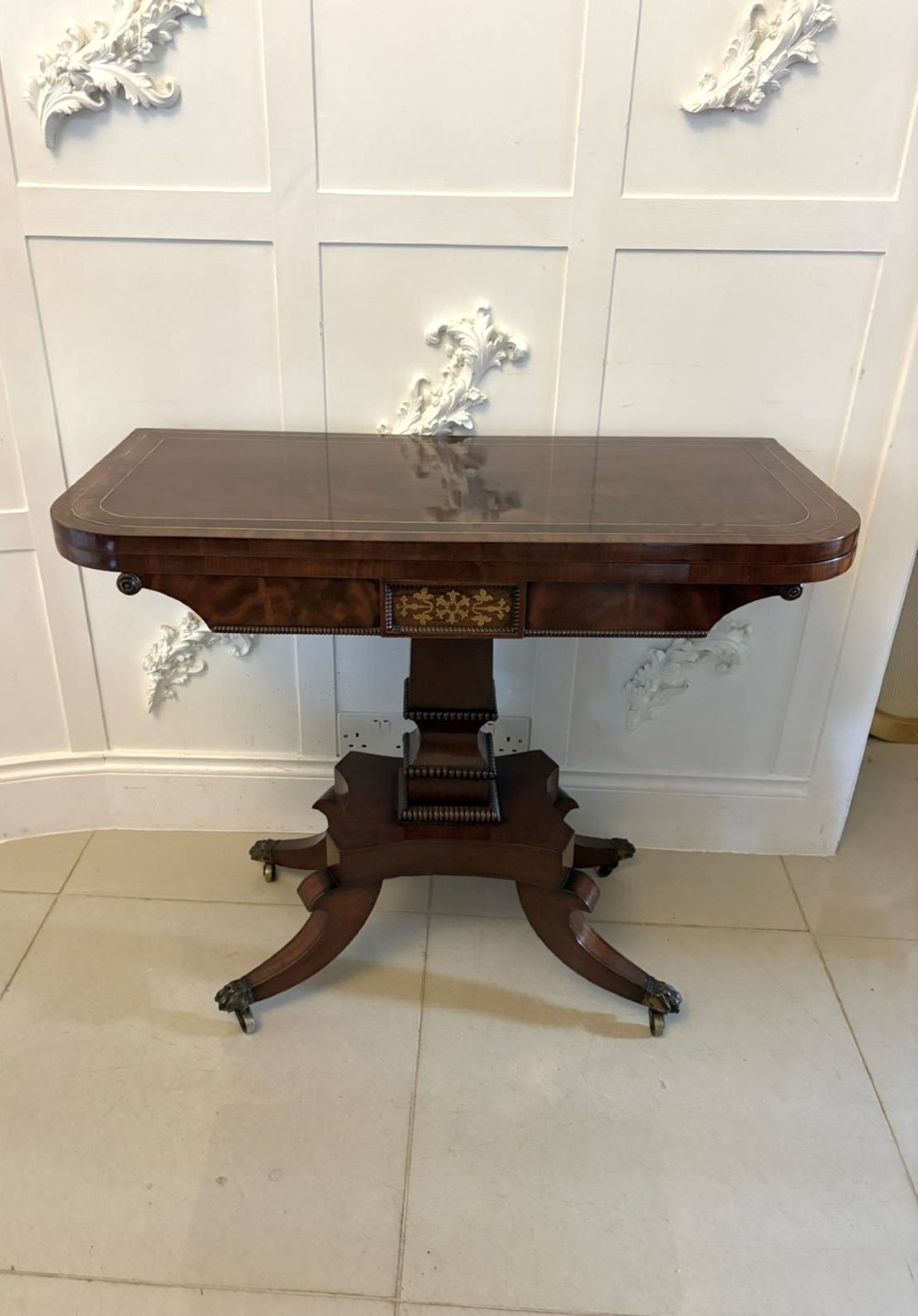An antique Regency mahogany fold-over card table featuring brass marquetry inlay, a central shaped pedestal, and a splayed base with brass castors.
