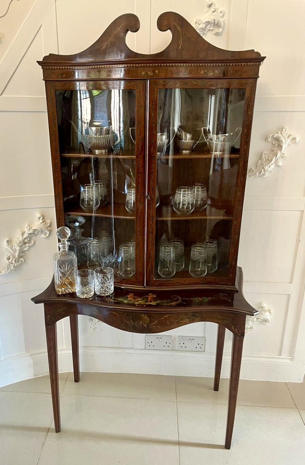 An antique mahogany display cabinet featuring a swan-neck pediment, glazed twin doors, hand-painted floral decoration, and slender tapered legs. View II