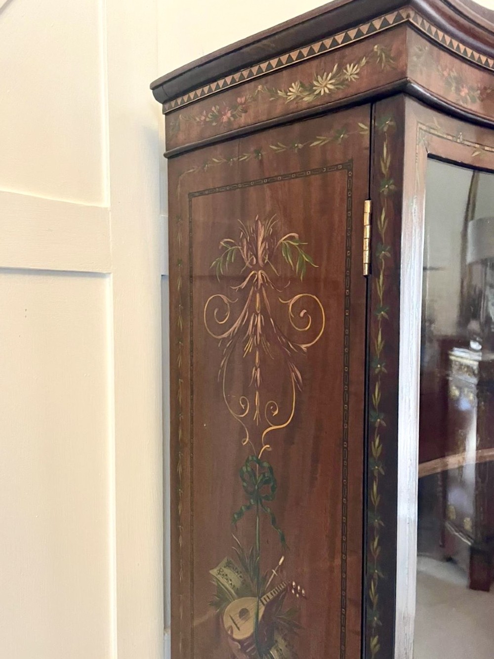 An antique mahogany display cabinet featuring a swan-neck pediment, glazed twin doors, hand-painted floral decoration, and slender tapered legs. View V