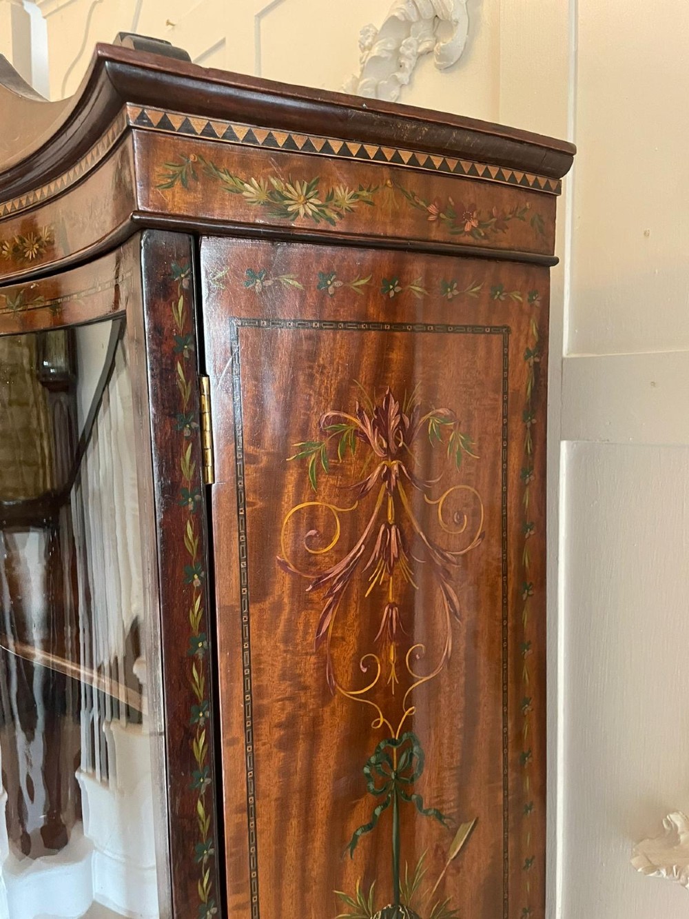 An antique mahogany display cabinet featuring a swan-neck pediment, glazed twin doors, hand-painted floral decoration, and slender tapered legs. View VI