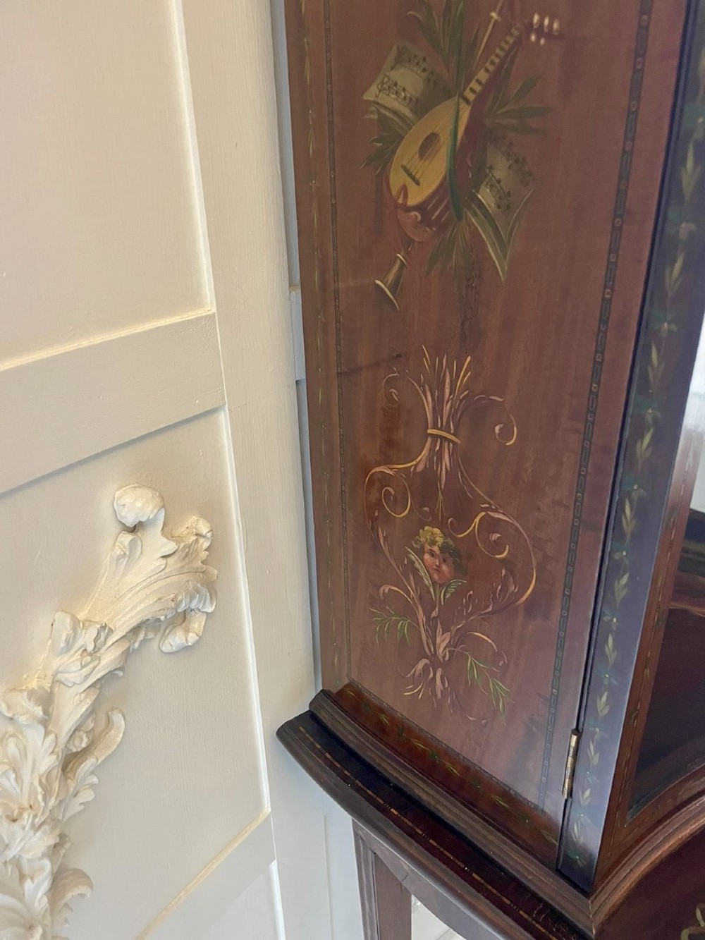 An antique mahogany display cabinet featuring a swan-neck pediment, glazed twin doors, hand-painted floral decoration, and slender tapered legs. View VII