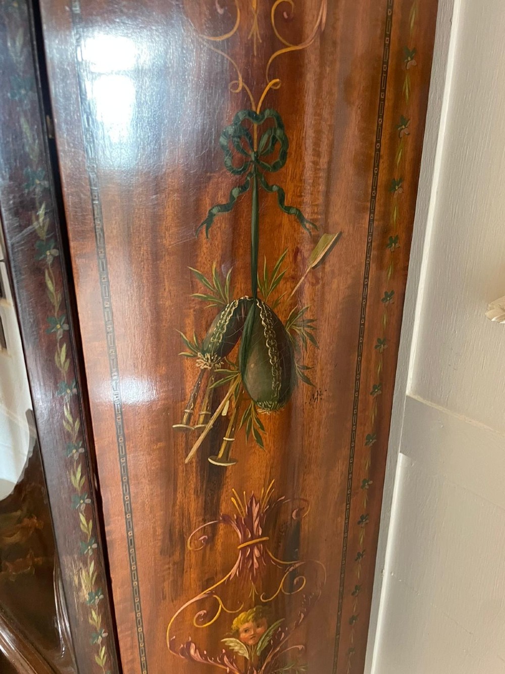 An antique mahogany display cabinet featuring a swan-neck pediment, glazed twin doors, hand-painted floral decoration, and slender tapered legs. View VIII