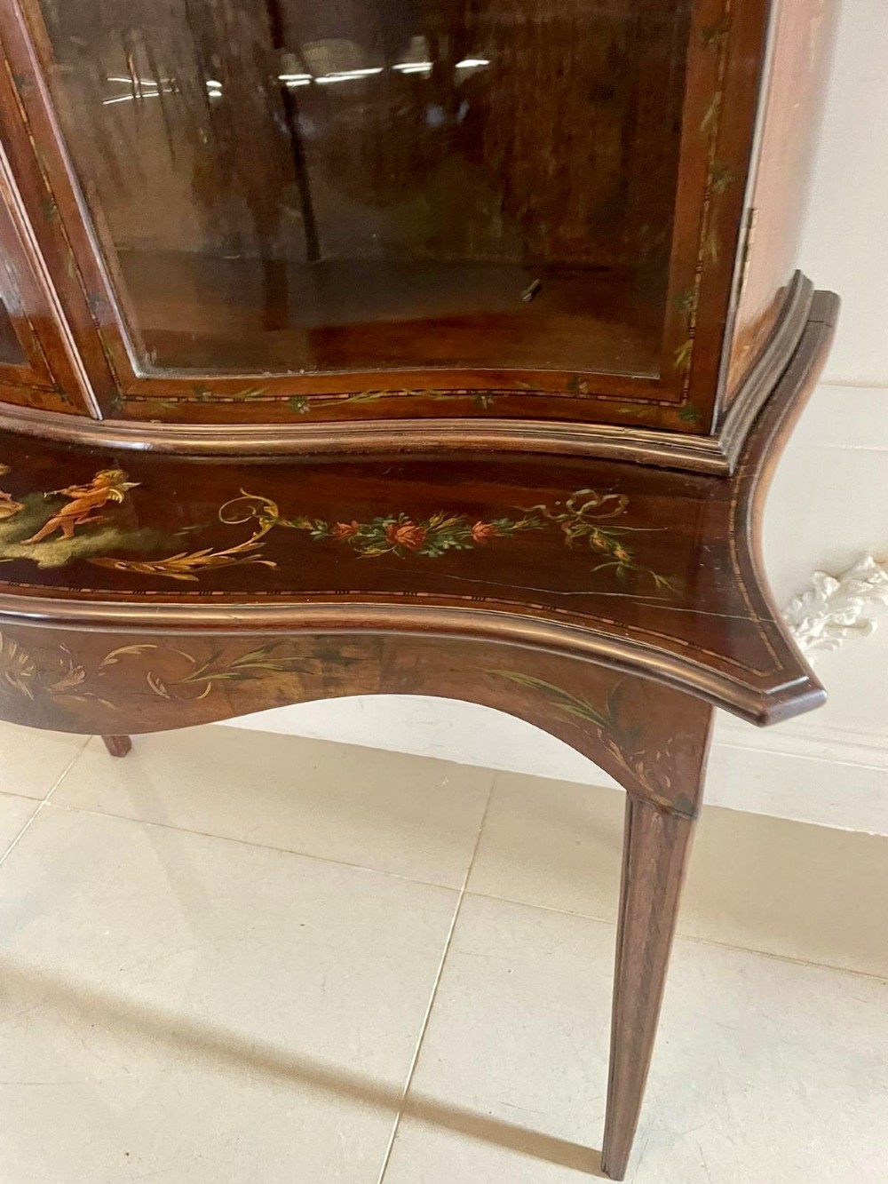 An antique mahogany display cabinet featuring a swan-neck pediment, glazed twin doors, hand-painted floral decoration, and slender tapered legs. View IX