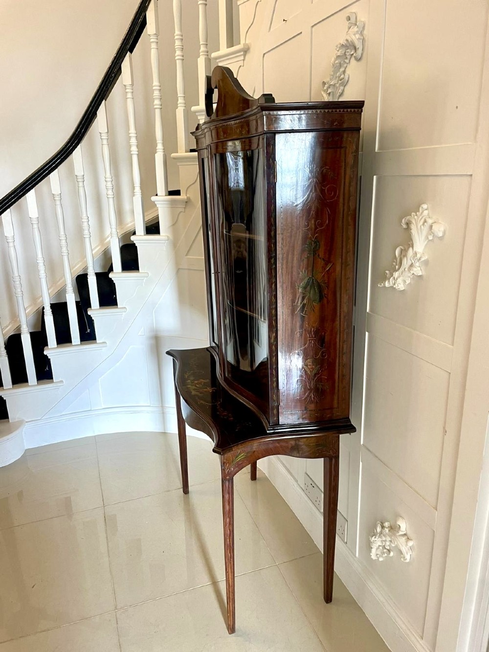 An antique mahogany display cabinet featuring a swan-neck pediment, glazed twin doors, hand-painted floral decoration, and slender tapered legs. View X