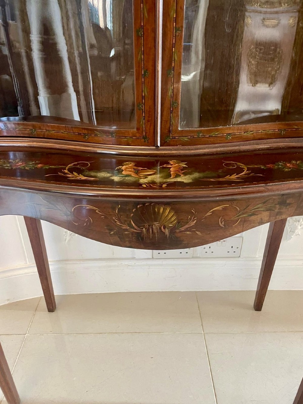 An antique mahogany display cabinet featuring a swan-neck pediment, glazed twin doors, hand-painted floral decoration, and slender tapered legs. View XI