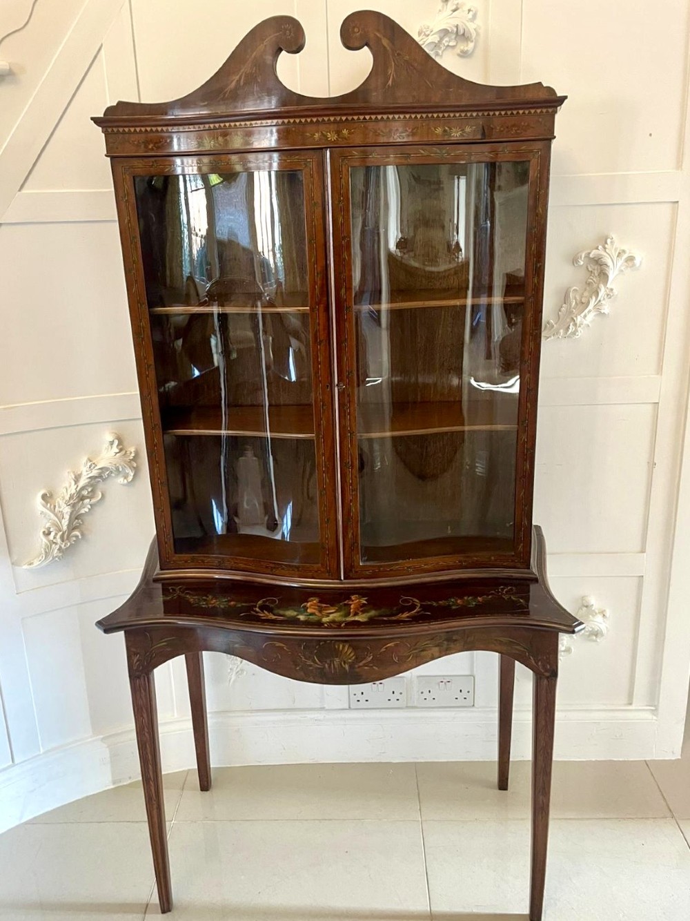 An antique mahogany display cabinet featuring a swan-neck pediment, glazed twin doors, hand-painted floral decoration, and slender tapered legs. View XV