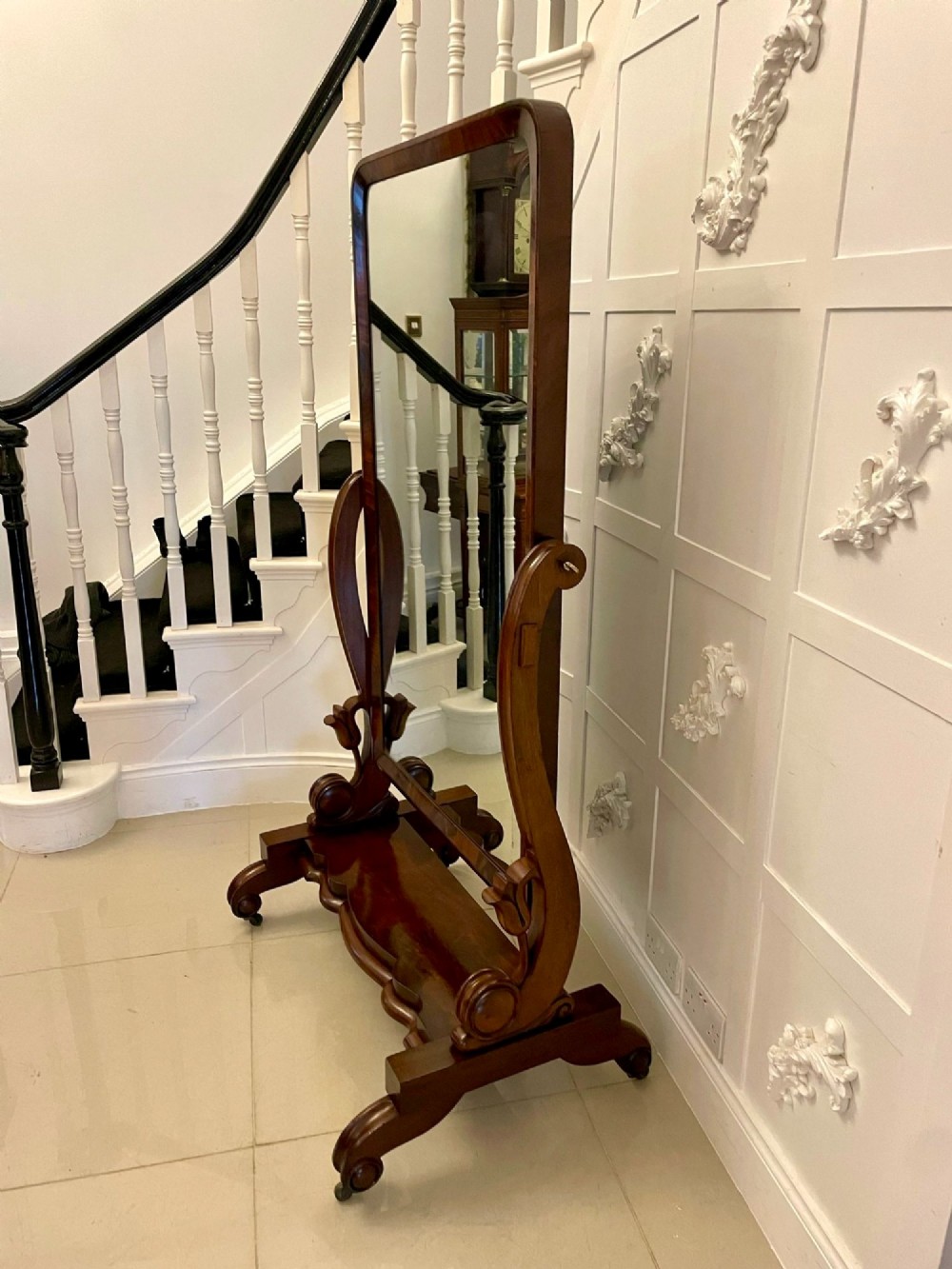 A full-length Victorian mahogany cheval mirror featuring a rounded rectangular swivelling frame, carved scroll uprights, a shaped plinth base, and brass castors. View IV