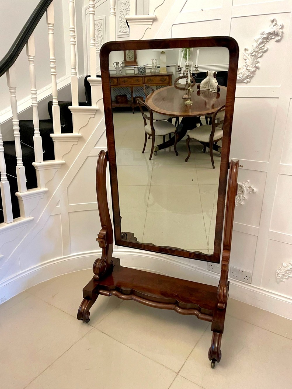 A full-length Victorian mahogany cheval mirror featuring a rounded rectangular swivelling frame, carved scroll uprights, a shaped plinth base, and brass castors. View V