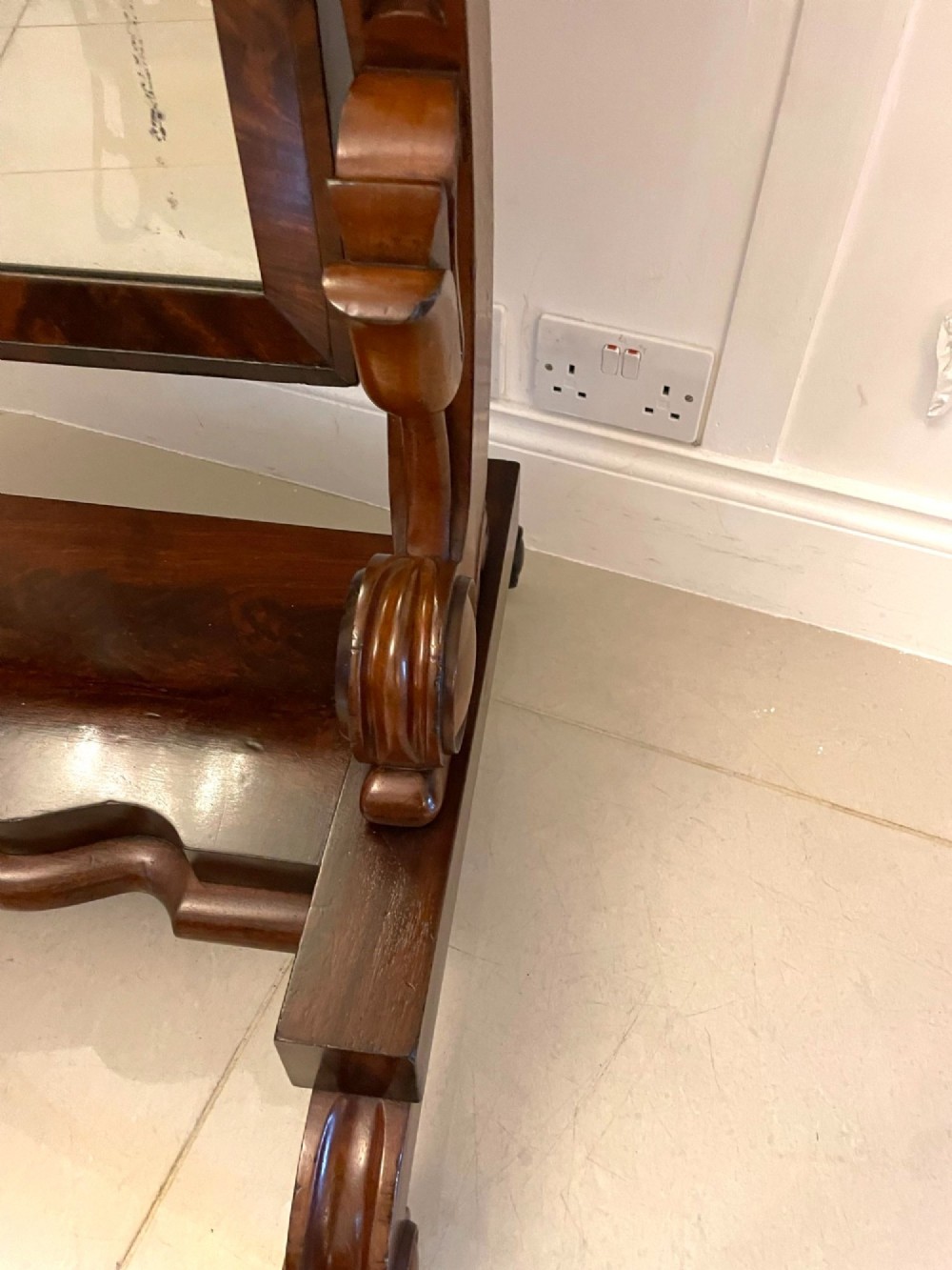 A full-length Victorian mahogany cheval mirror featuring a rounded rectangular swivelling frame, carved scroll uprights, a shaped plinth base, and brass castors. View VI