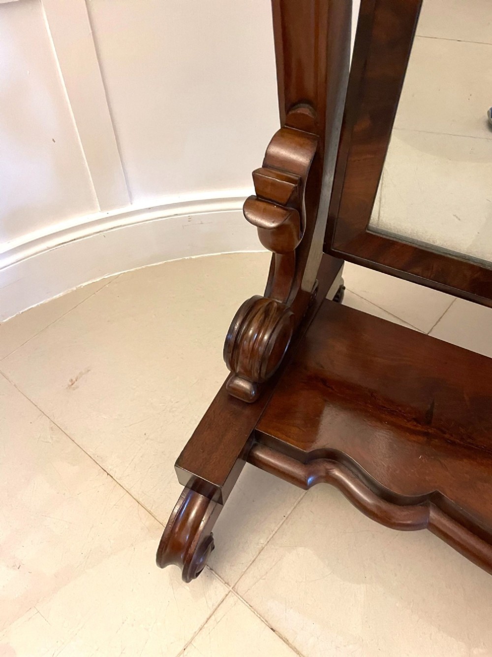 A full-length Victorian mahogany cheval mirror featuring a rounded rectangular swivelling frame, carved scroll uprights, a shaped plinth base, and brass castors. View VII