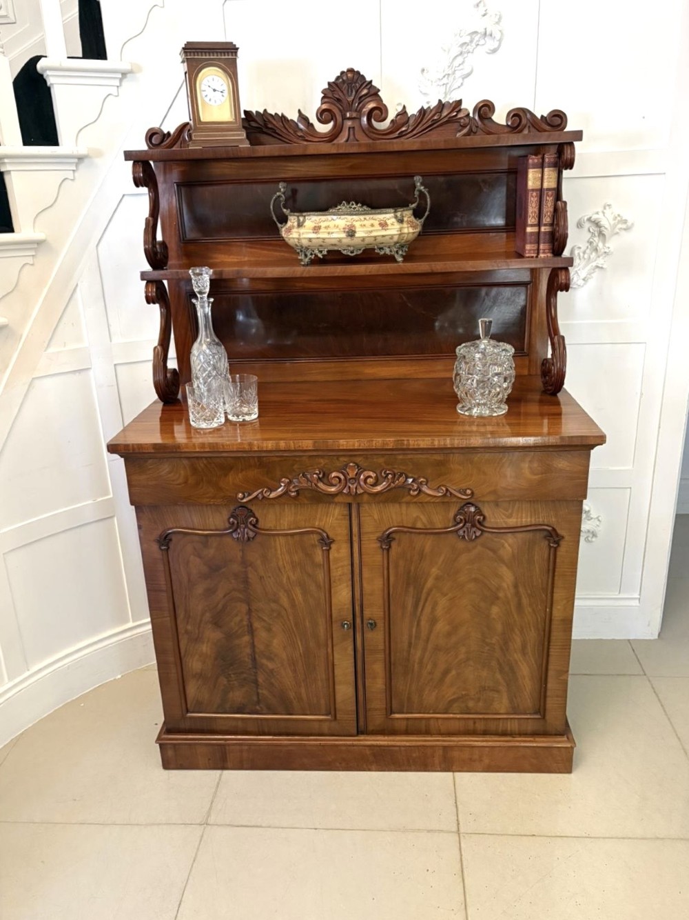 A Victorian mahogany chiffonier featuring a two-tiered upper shelf superstructure with carved scroll supports and a lower cupboard with flame mahogany veneered doors. View IV