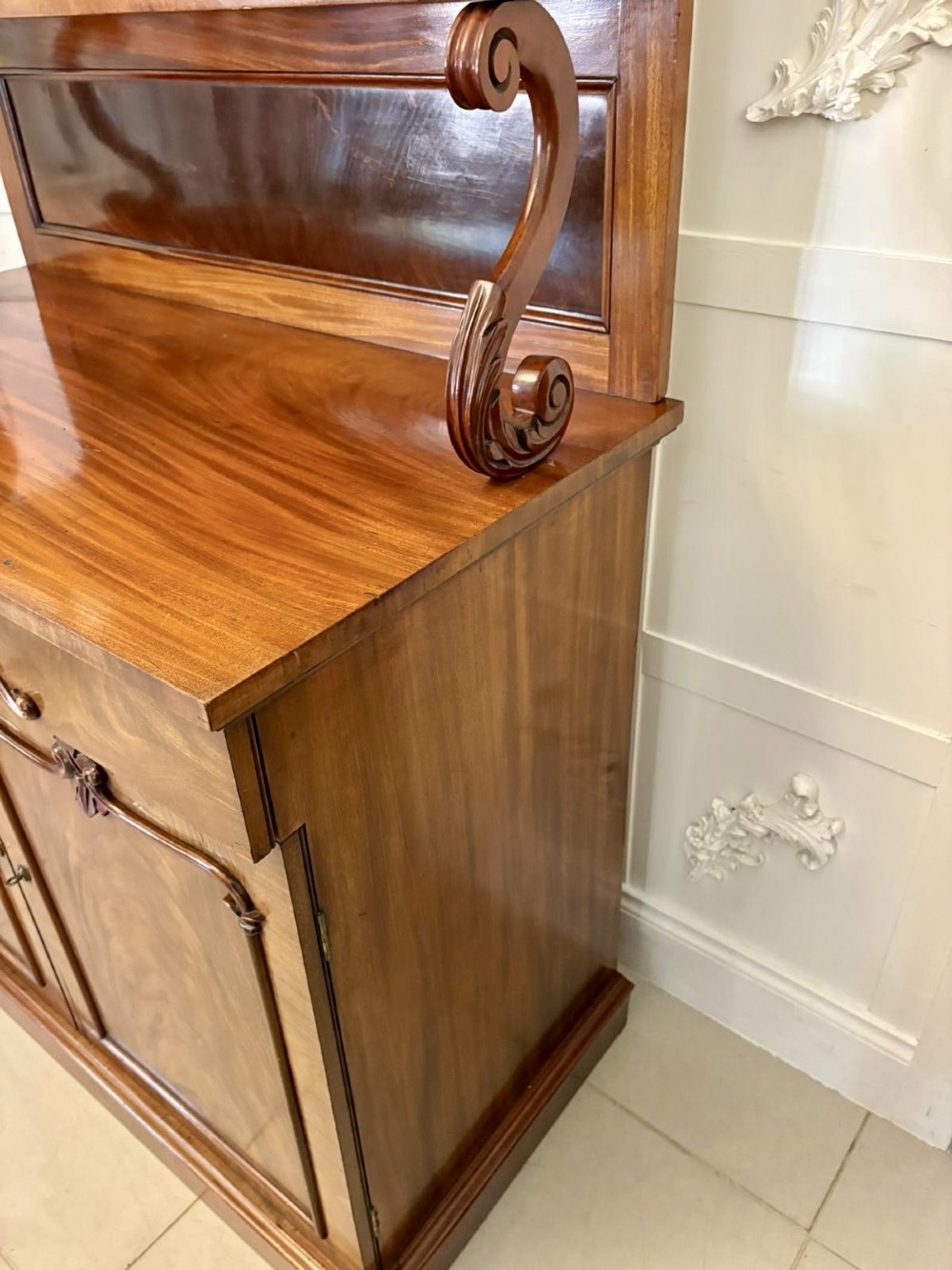 A Victorian mahogany chiffonier featuring a two-tiered upper shelf superstructure with carved scroll supports and a lower cupboard with flame mahogany veneered doors. View X