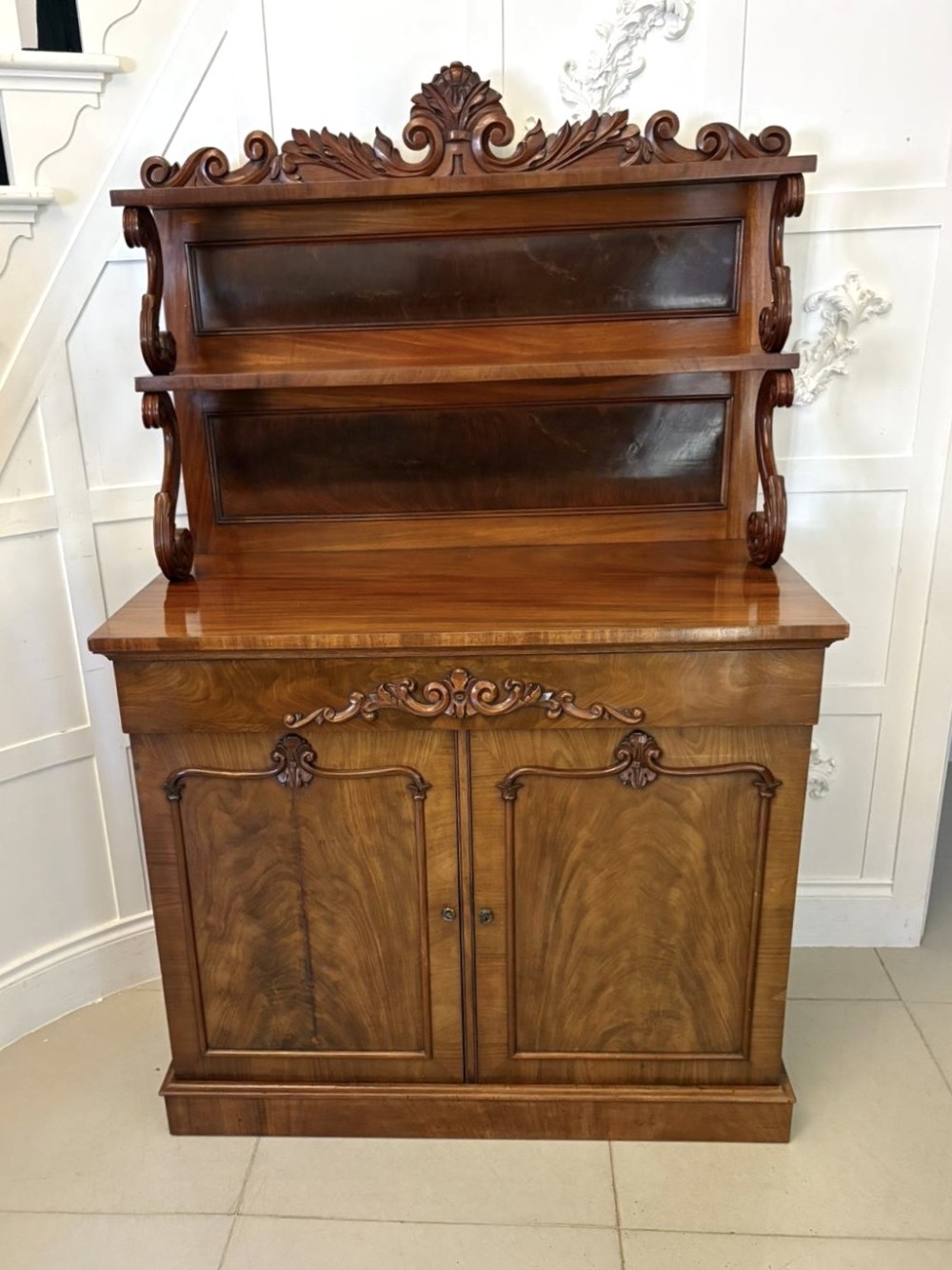 A Victorian mahogany chiffonier featuring a two-tiered upper shelf superstructure with carved scroll supports and a lower cupboard with flame mahogany veneered doors.