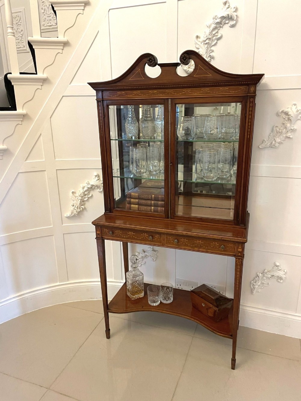 An antique mahogany display cabinet featuring a swan neck pediment, marquetry inlays, glazed double doors, interior glass shelves, and square tapered legs. View III
