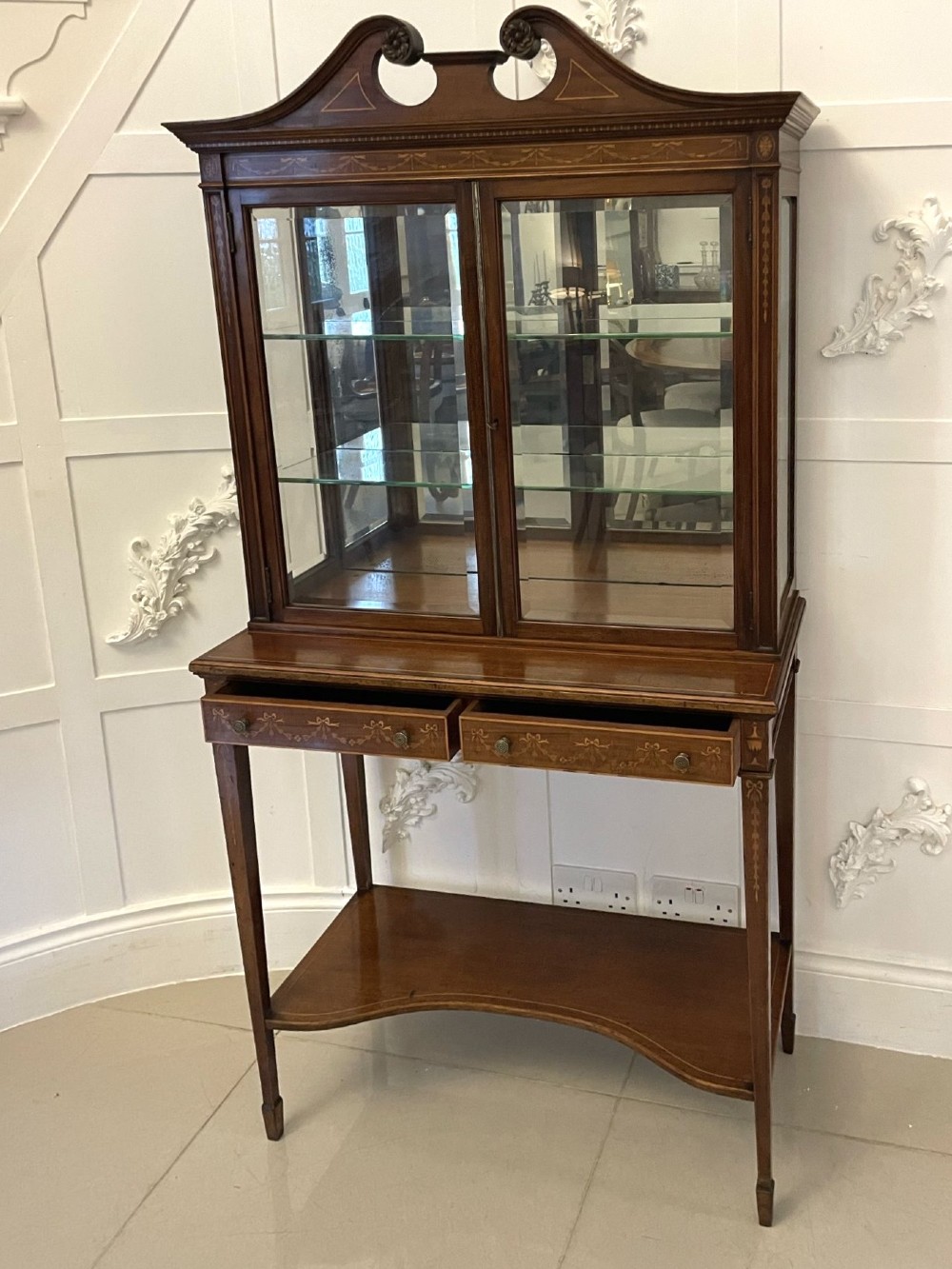 An antique mahogany display cabinet featuring a swan neck pediment, marquetry inlays, glazed double doors, interior glass shelves, and square tapered legs. View VI