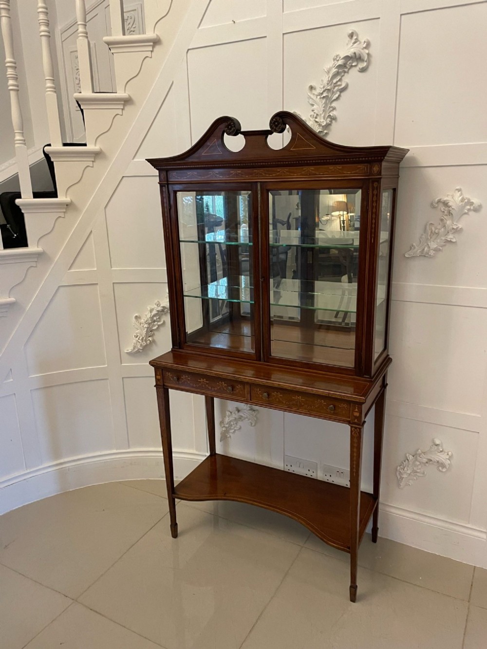 An antique mahogany display cabinet featuring a swan neck pediment, marquetry inlays, glazed double doors, interior glass shelves, and square tapered legs. View XV