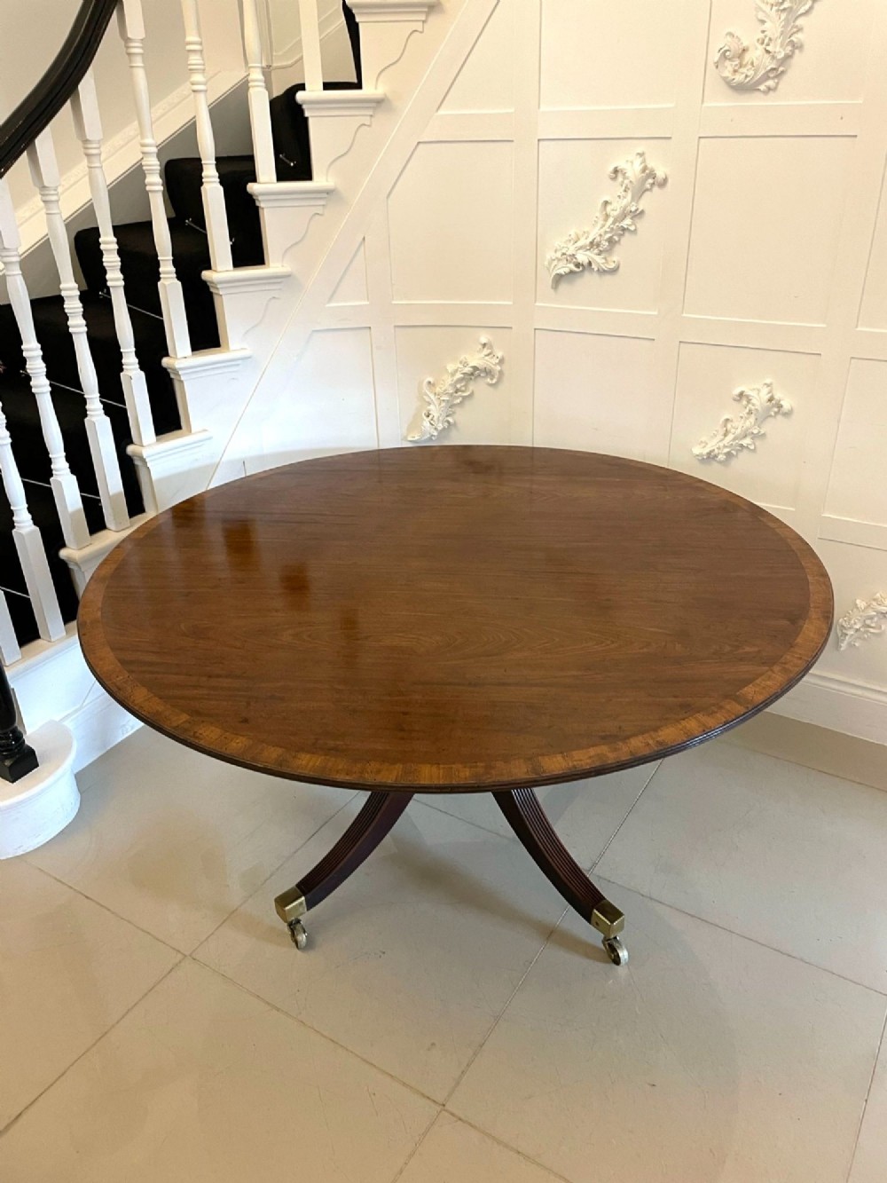 A circular George III figured mahogany tilt-top dining table positioned vertically on a four-legged pedestal base with brass castors. View IV