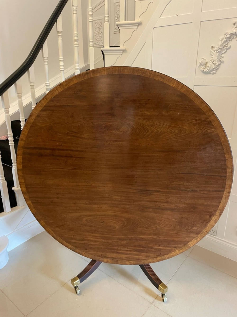 A circular George III figured mahogany tilt-top dining table positioned vertically on a four-legged pedestal base with brass castors. View IX