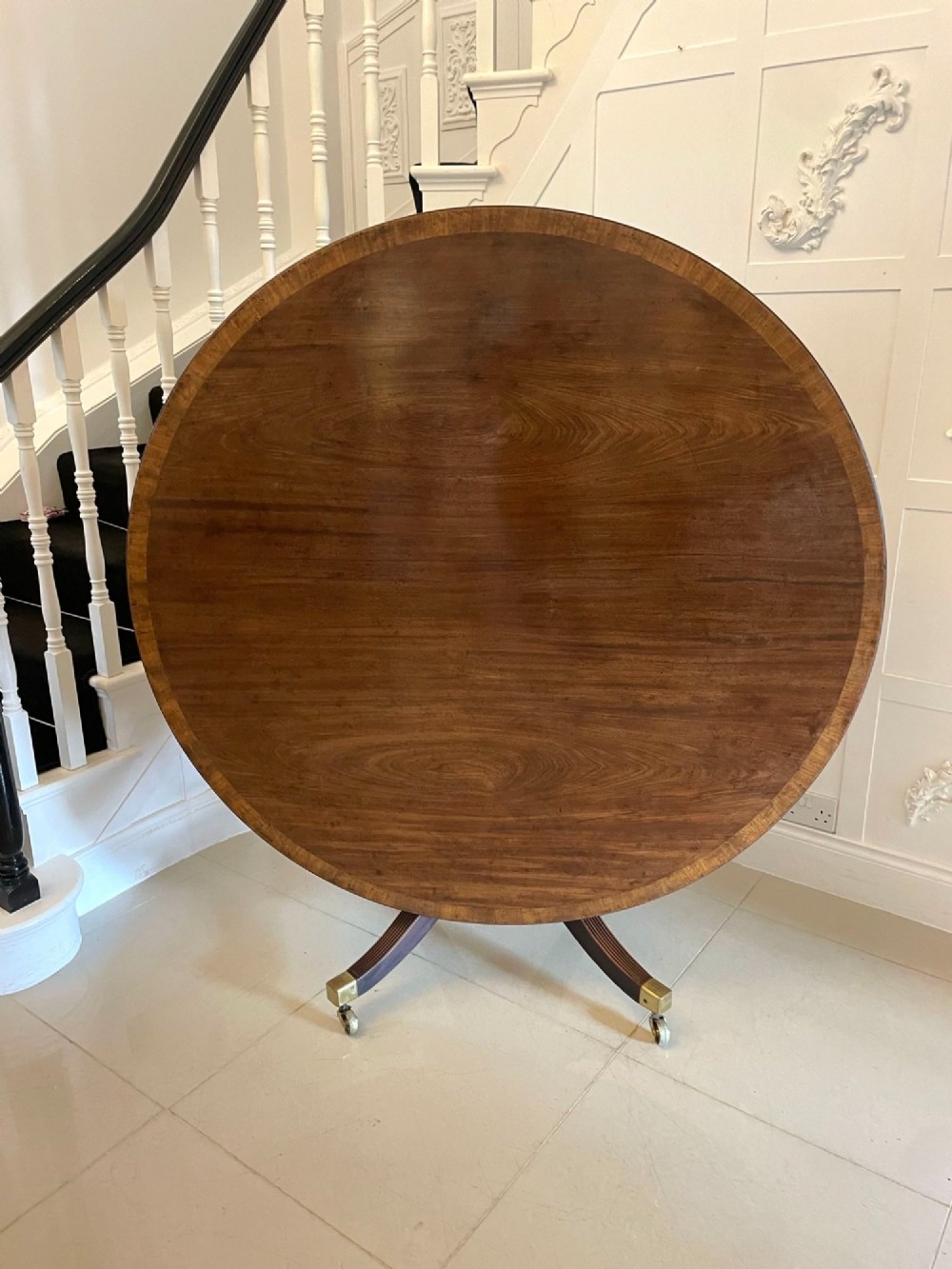 A circular George III figured mahogany tilt-top dining table positioned vertically on a four-legged pedestal base with brass castors. View X