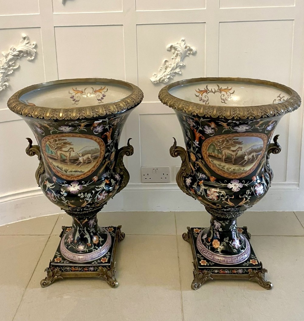 A pair of black French porcelain jardiniere urns featuring polychrome floral enamel, central painted animal reserves, and gadrooned brass mounts on square plinths.