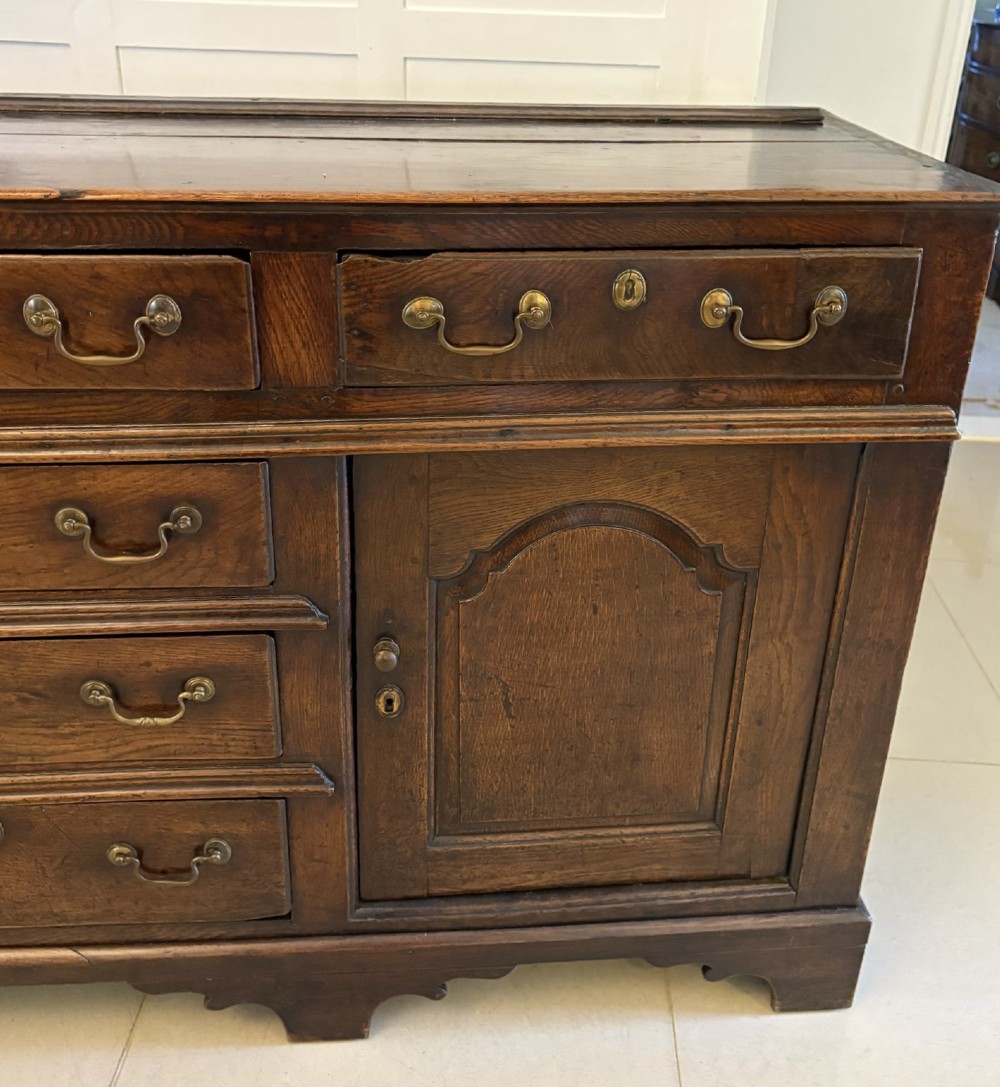 A George III dark oak dresser base featuring multiple drawers, arched cupboard doors, and brass swan-neck handles. View IV